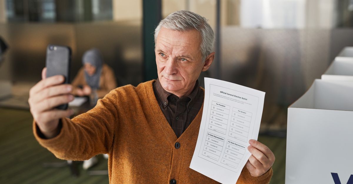 Elderly man with gray hair holding a ballot and taking a selfie in a voting booth.